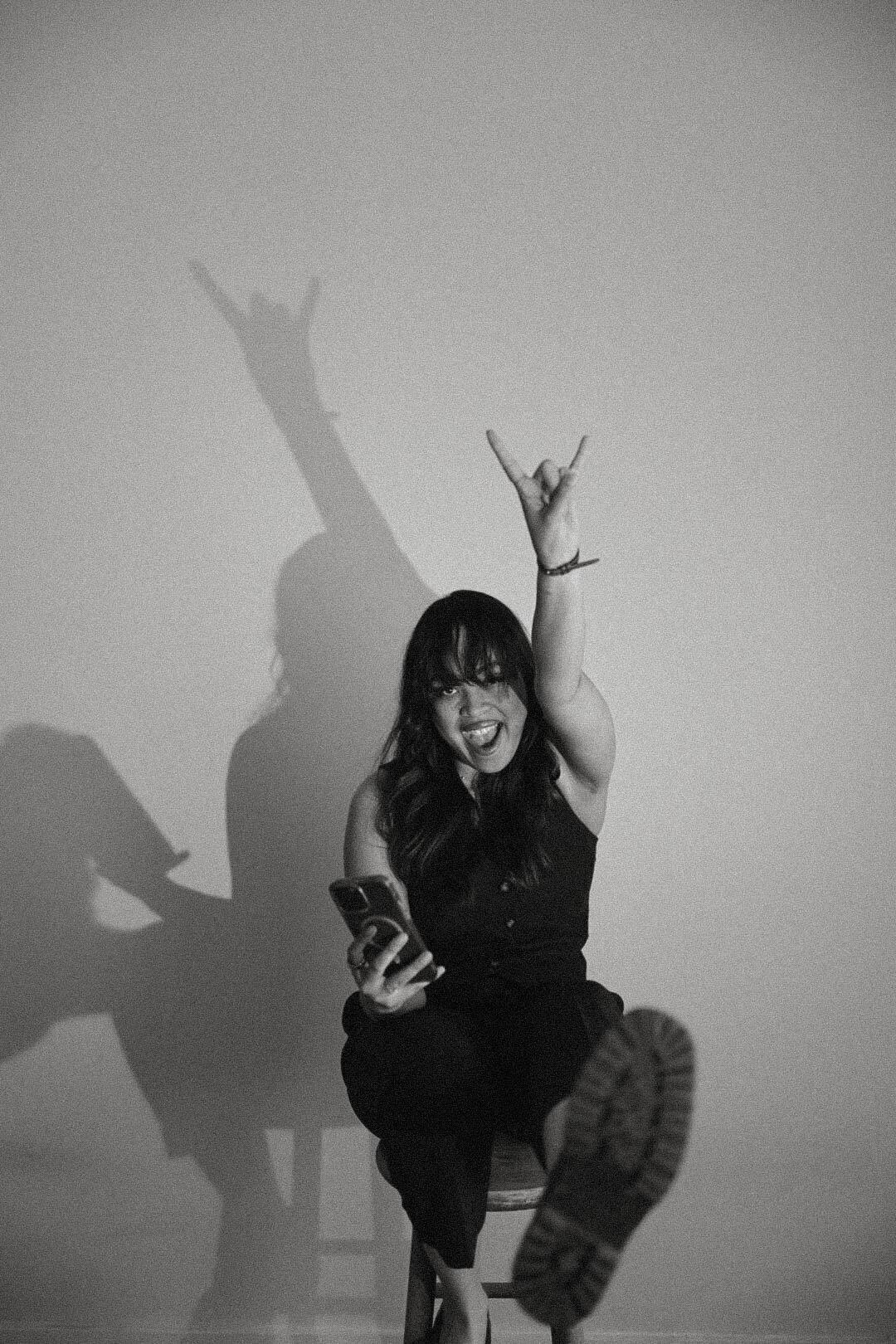 Hallel Saine seated on a studio stool, arm raised in horns gesture, dramatic cast shadow